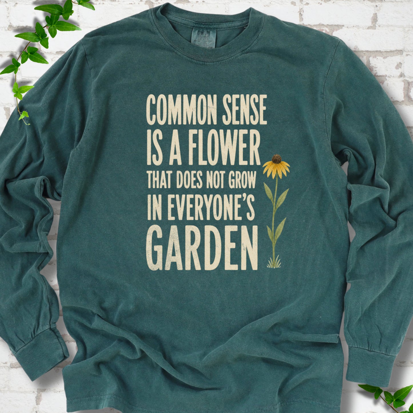 Blue Spruce Comfort Colors® long sleeve T-shirt flat lay with cream text reading "Common sense is a flower that does not grow in everyone's garden" beside a yellow flower