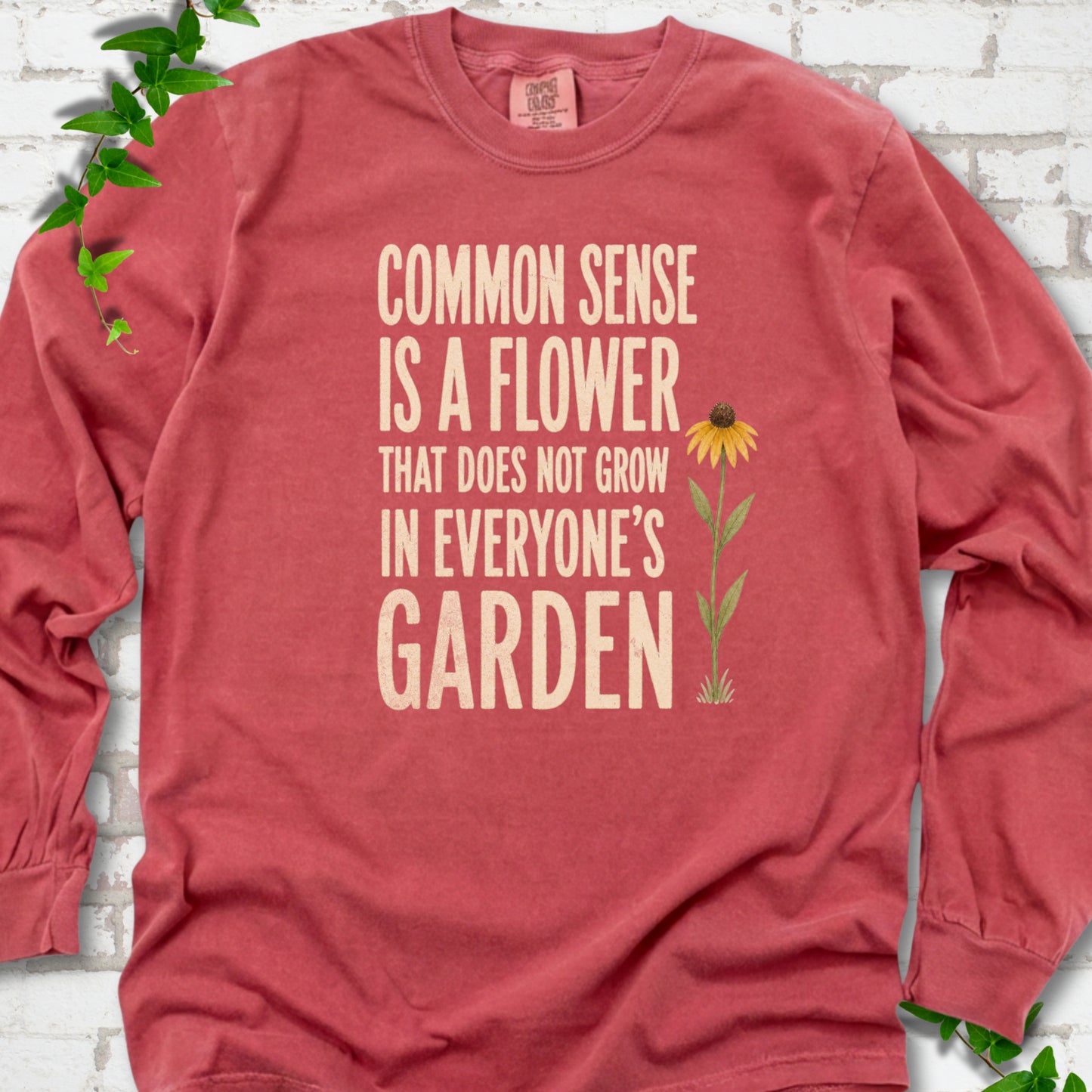 Crimson Comfort Colors® long sleeve T-shirt flat lay with cream text reading "Common sense is a flower that does not grow in everyone's garden" beside a yellow flower