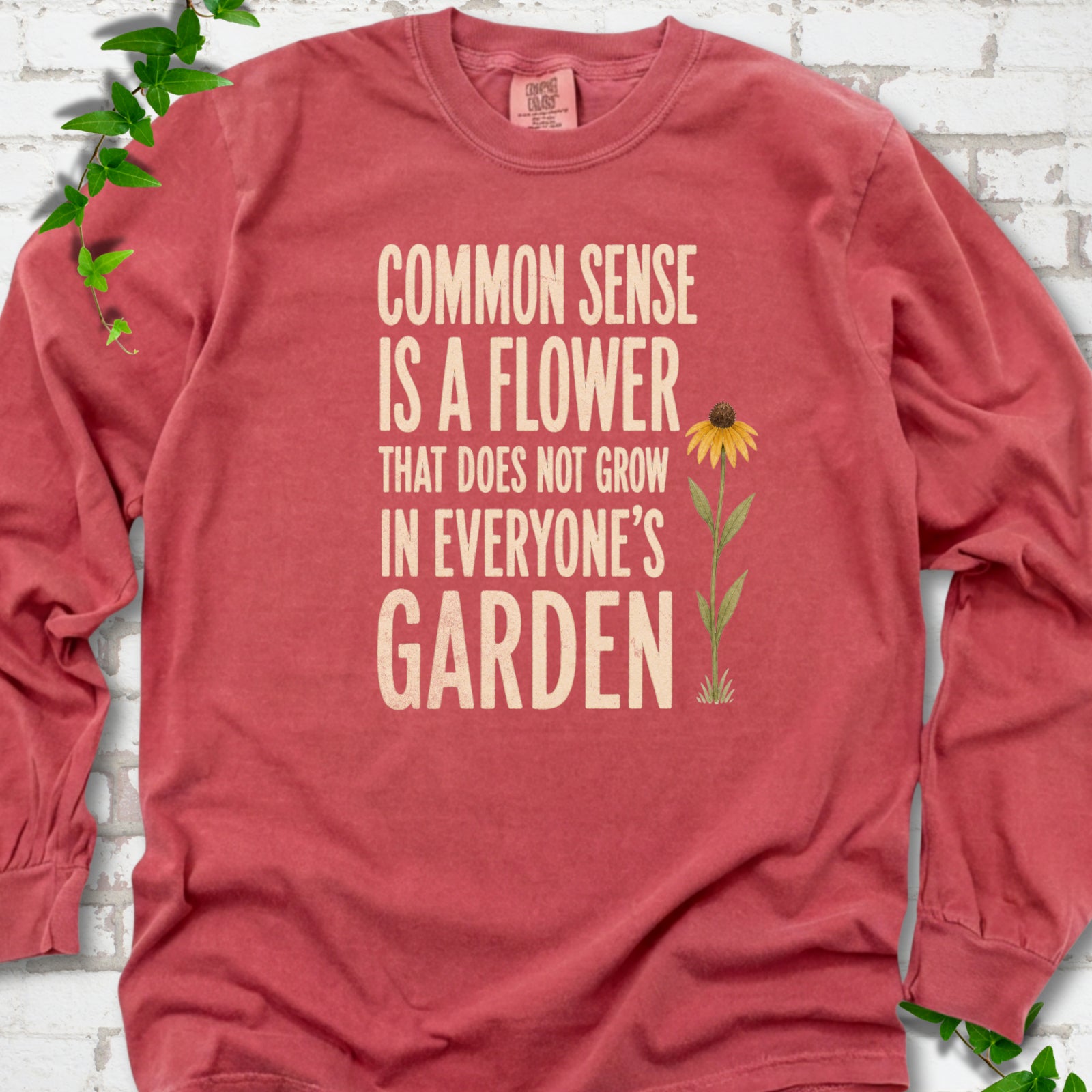 Crimson Comfort Colors® long sleeve T-shirt flat lay with cream text reading "Common sense is a flower that does not grow in everyone's garden" beside a yellow flower