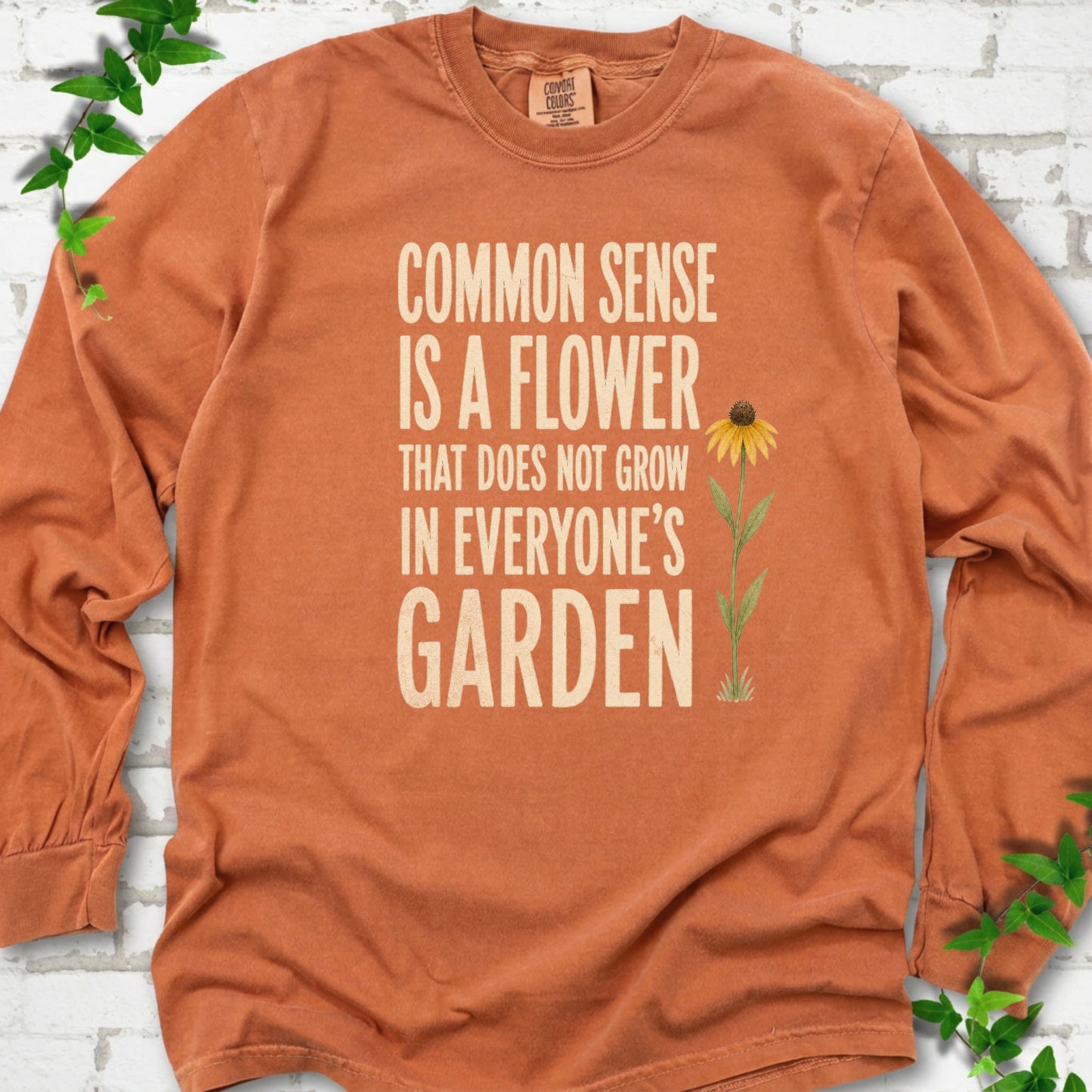 Yam Comfort Colors® long sleeve T-shirt flat lay with cream text reading "Common sense is a flower that does not grow in everyone's garden" beside a yellow flower