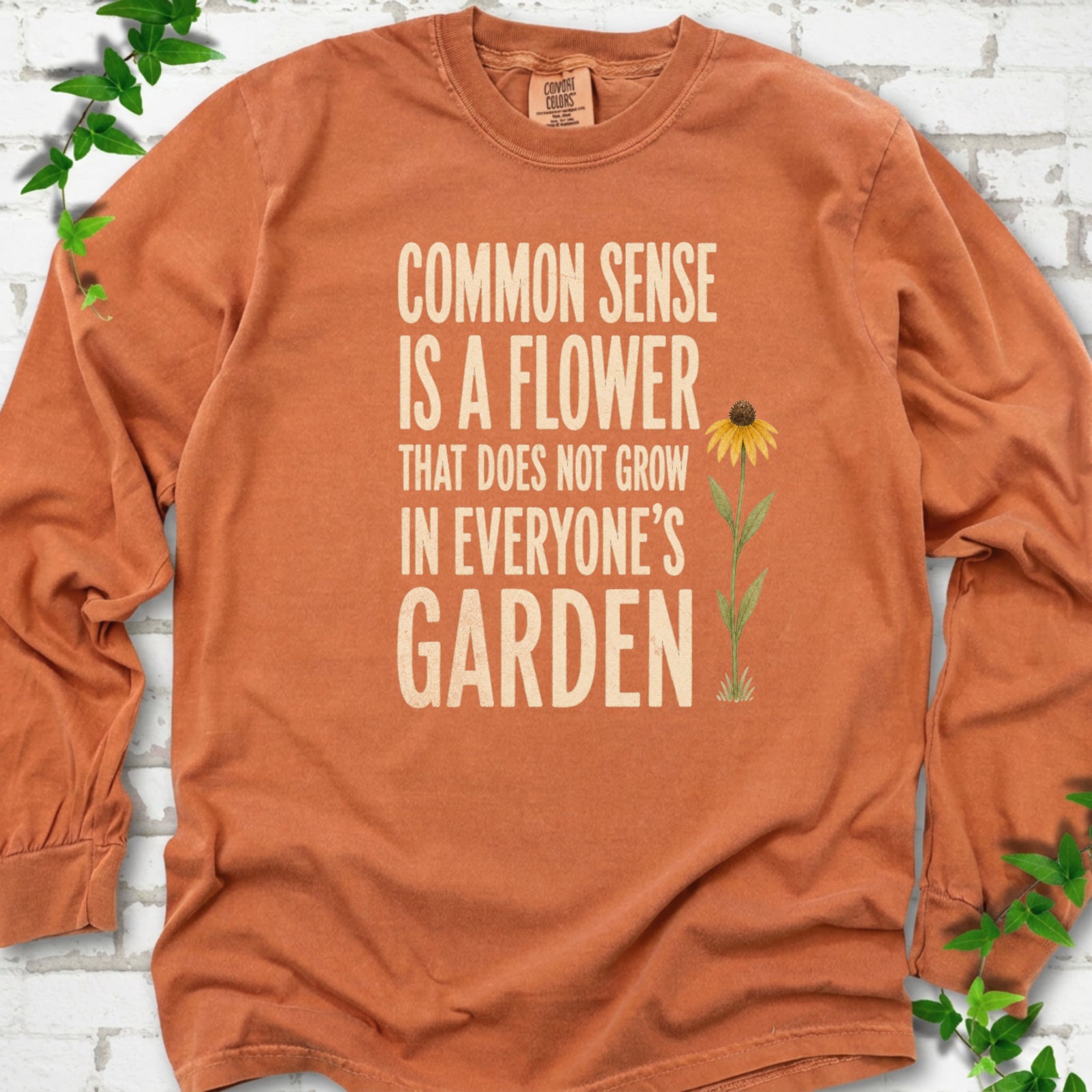 Yam Comfort Colors® long sleeve T-shirt flat lay with cream text reading "Common sense is a flower that does not grow in everyone's garden" beside a yellow flower