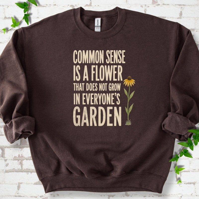 Dark Chocolate Gildan® 18000 crewneck sweatshirt flat lay with cream text reading ‘Common sense is a flower that does not grow in everyone's garden’ and a yellow flower