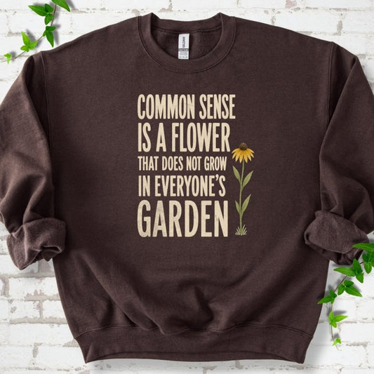 Dark Chocolate Gildan® 18000 crewneck sweatshirt flat lay with cream text reading ‘Common sense is a flower that does not grow in everyone's garden’ and a yellow flower