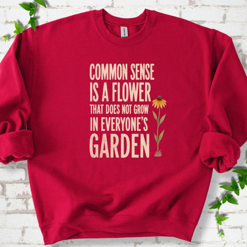 Cardinal Red Gildan® 18000 crewneck sweatshirt flat lay with cream text reading ‘Common sense is a flower that does not grow in everyone's garden’ and a yellow flower