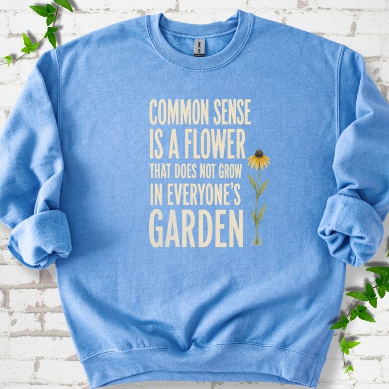 Carolina Blue Gildan® 18000 crewneck sweatshirt flat lay with cream text reading ‘Common sense is a flower that does not grow in everyone's garden’ and a yellow flower