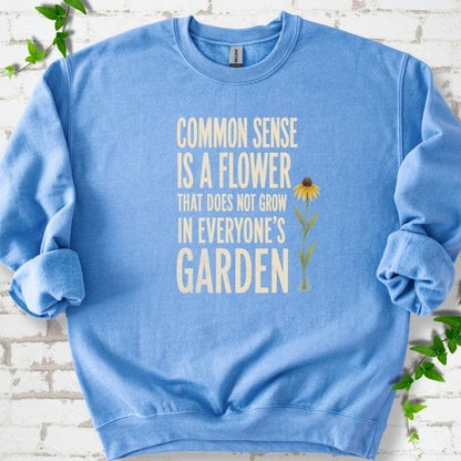 Carolina Blue Gildan® 18000 crewneck sweatshirt flat lay with cream text reading ‘Common sense is a flower that does not grow in everyone's garden’ and a yellow flower