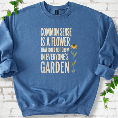 Indigo Blue Gildan® 18000 crewneck sweatshirt flat lay with cream text reading ‘Common sense is a flower that does not grow in everyone's garden’ and a yellow flower