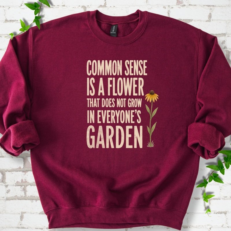 Maroon Gildan® 18000 crewneck sweatshirt flat lay with cream text reading ‘Common sense is a flower that does not grow in everyone's garden’ and a yellow flower