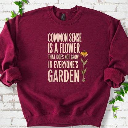 Maroon Gildan® 18000 crewneck sweatshirt flat lay with cream text reading ‘Common sense is a flower that does not grow in everyone's garden’ and a yellow flower