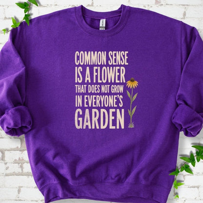 Purple Gildan® 18000 crewneck sweatshirt flat lay with cream text reading ‘Common sense is a flower that does not grow in everyone's garden’ and a yellow flower