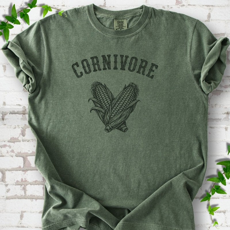 Moss Comfort Colors® short sleeve T-shirt flat lay featuring black line art of two sweet corn cobs beneath the word "Cornivore"