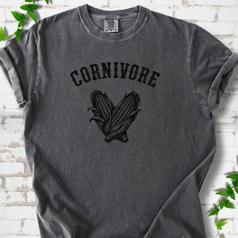 Pepper Comfort Colors® short sleeve T-shirt flat lay featuring black line art of two sweet corn cobs beneath the word "Cornivore"