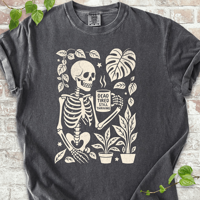 Dead Tired T-shirt on Comfort Colors™ Pepper – shows a tired skeleton graphic holding a coffee cup and surrounded by plants, Monstera leaves, and stars