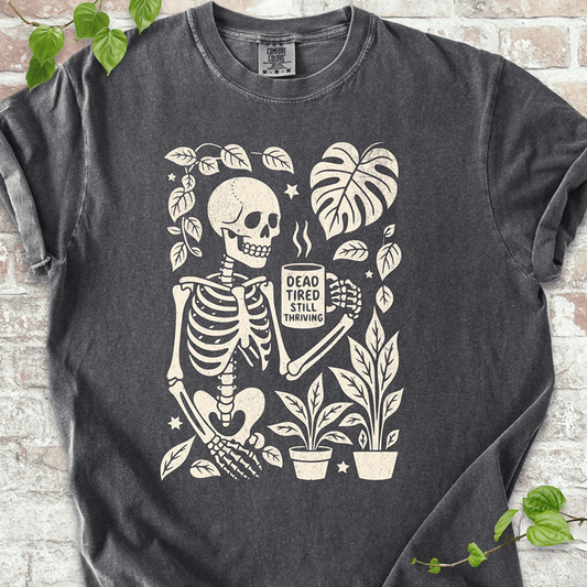 Dead Tired T-shirt on Comfort Colors™ Pepper – shows a tired skeleton graphic holding a coffee cup and surrounded by plants, Monstera leaves, and stars