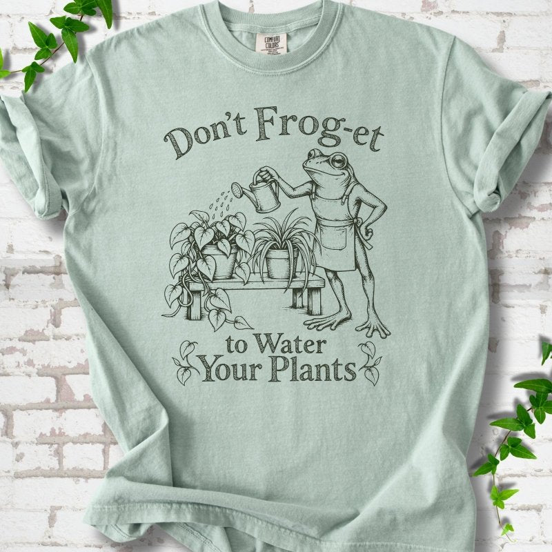 Bay Comfort Colors® T-shirt laid flat, showing a frog in an apron watering potted plants, with “Don’t Frog-et to Water Your Plants” lettering.