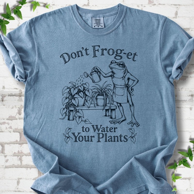 Blue Jean Comfort Colors® T-shirt laid flat, showing a frog in an apron watering potted plants, with “Don’t Frog-et to Water Your Plants” lettering.