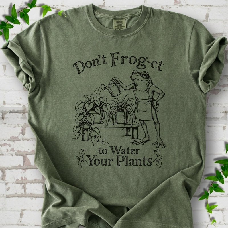 Moss Comfort Colors® T-shirt laid flat, showing a frog in an apron watering potted plants, with “Don’t Frog-et to Water Your Plants” lettering.