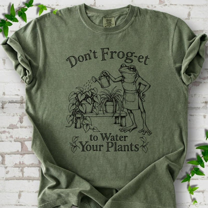 Moss Comfort Colors® T-shirt laid flat, showing a frog in an apron watering potted plants, with “Don’t Frog-et to Water Your Plants” lettering.
