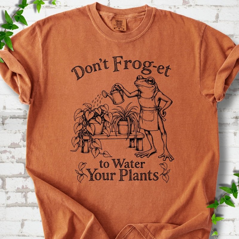 Yam Comfort Colors® T-shirt laid flat, showing a frog in an apron watering potted plants, with “Don’t Frog-et to Water Your Plants” lettering.