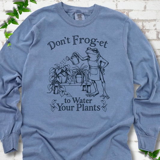 Blue Jean Comfort Colors® long sleeve T-shirt laid flat, showing a frog in an apron watering potted plants, with “Don’t Frog-et to Water Your Plants” lettering.