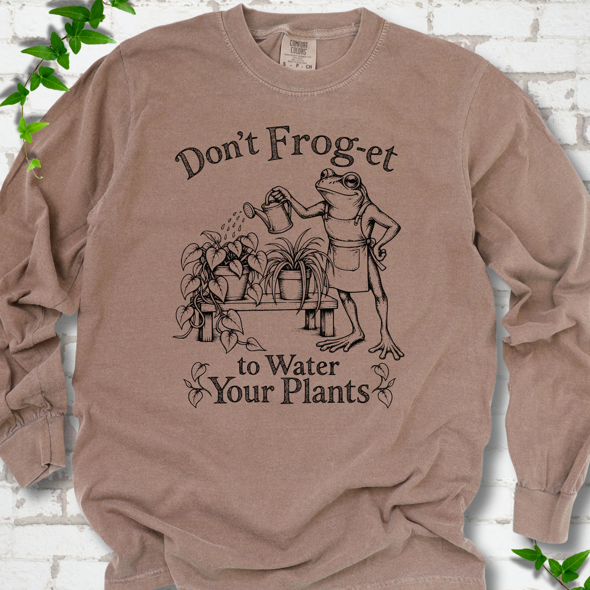 Espresso Comfort Colors® long sleeve T-shirt laid flat, showing a frog in an apron watering potted plants, with “Don’t Frog-et to Water Your Plants” lettering.