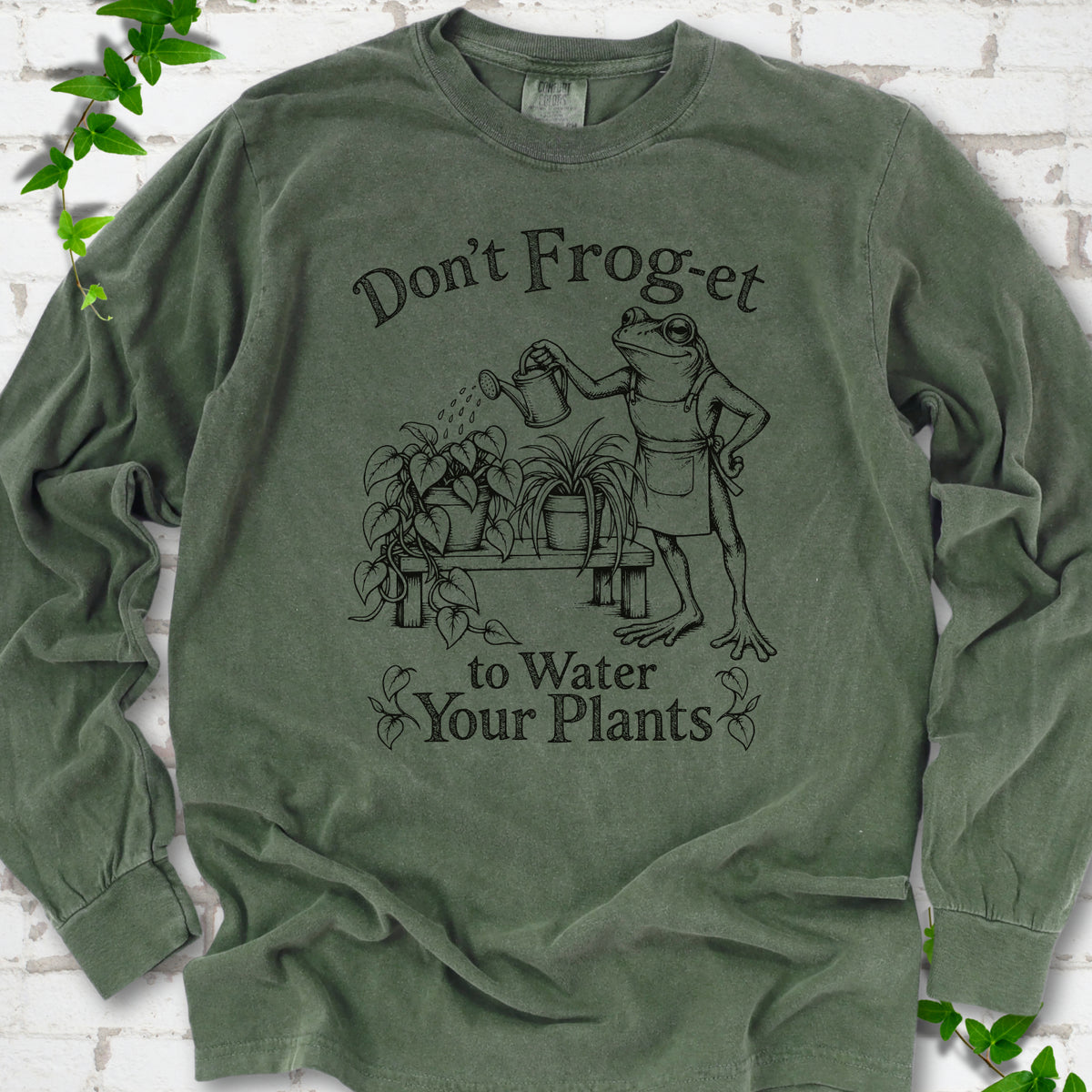 Hemp Comfort Colors® long sleeve T-shirt laid flat, showing a frog in an apron watering potted plants, with “Don’t Frog-et to Water Your Plants” lettering.