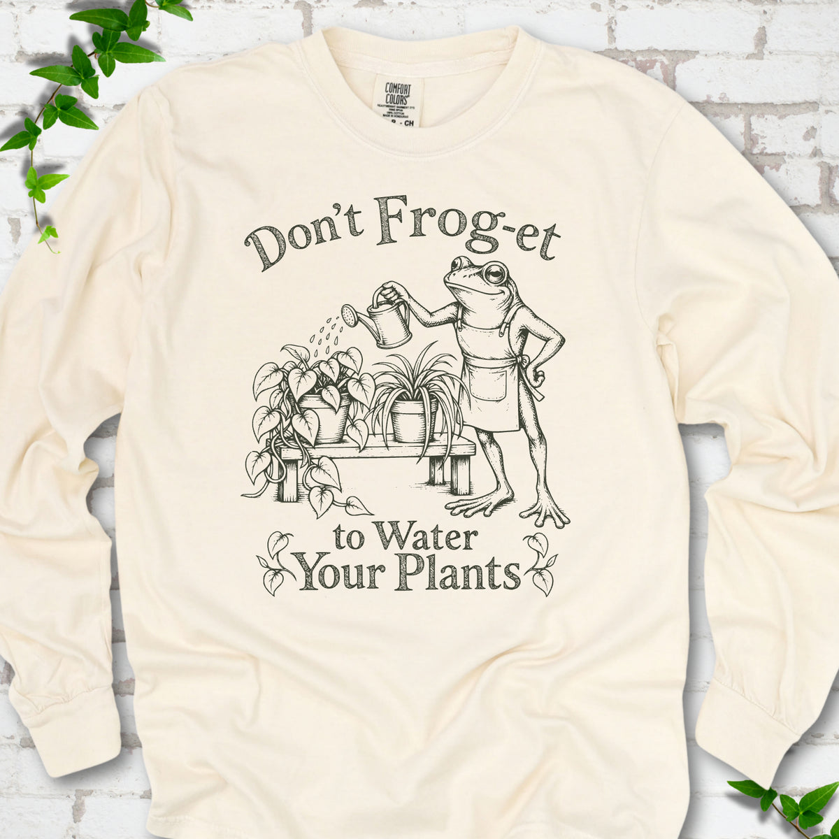Ivory Comfort Colors® long sleeve T-shirt laid flat, showing a frog in an apron watering potted plants, with “Don’t Frog-et to Water Your Plants” lettering.