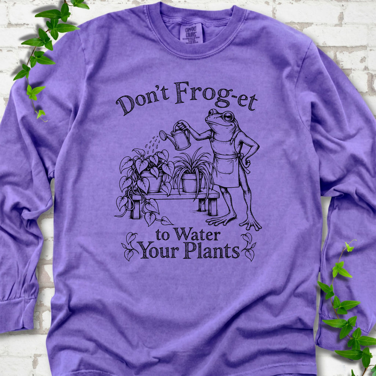 Violet Comfort Colors® long sleeve T-shirt laid flat, showing a frog in an apron watering potted plants, with “Don’t Frog-et to Water Your Plants” lettering.