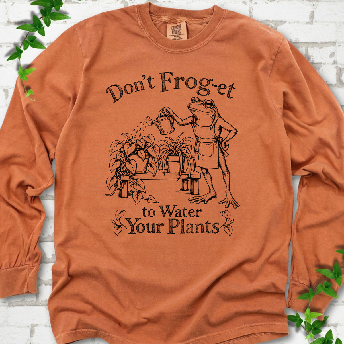 Yam Comfort Colors® long sleeve T-shirt laid flat, showing a frog in an apron watering potted plants, with “Don’t Frog-et to Water Your Plants” lettering.