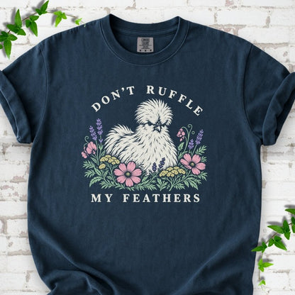 Dark navy T-shirt flat lay with a fluffy white Silkie hen with a fierce scowl sitting among pink cosmos, lavender, sweet pea and yellow yarrow, with "Don't Ruffle" arched above and "My Feathers" below, on white brick with ivy.