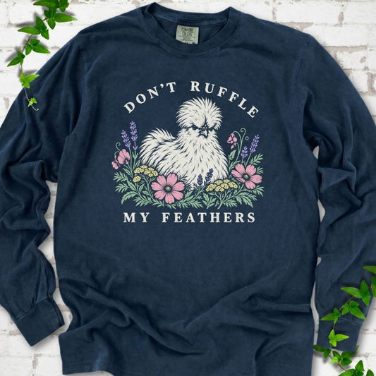 Dark navy long sleeve T-shirt flat lay with a fluffy white Silkie hen with a fierce scowl sitting among pink cosmos, lavender, sweet pea and yellow yarrow, with "Don't Ruffle" arched above and "My Feathers" below, on white brick with ivy.