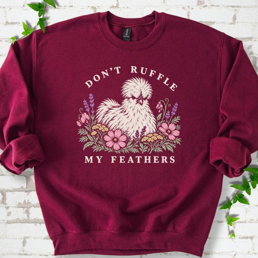 Deep maroon sweatshirt flat lay with a fluffy white Silkie hen with a fierce scowl sitting among pink cosmos, lavender, sweet pea and yellow yarrow, with "Don't Ruffle" arched above and "My Feathers" below, on white brick with ivy.