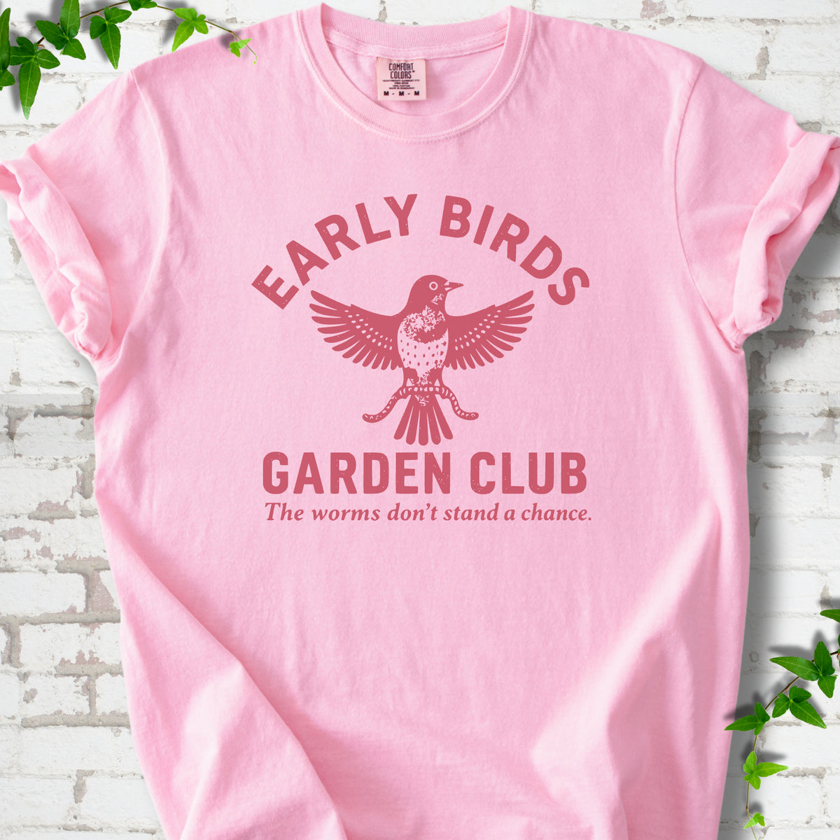 Blossom short sleeve T-shirt laid flat on white brick with greenery. Red bird holding worms between "Early Birds" and "Garden Club" chest lettering.