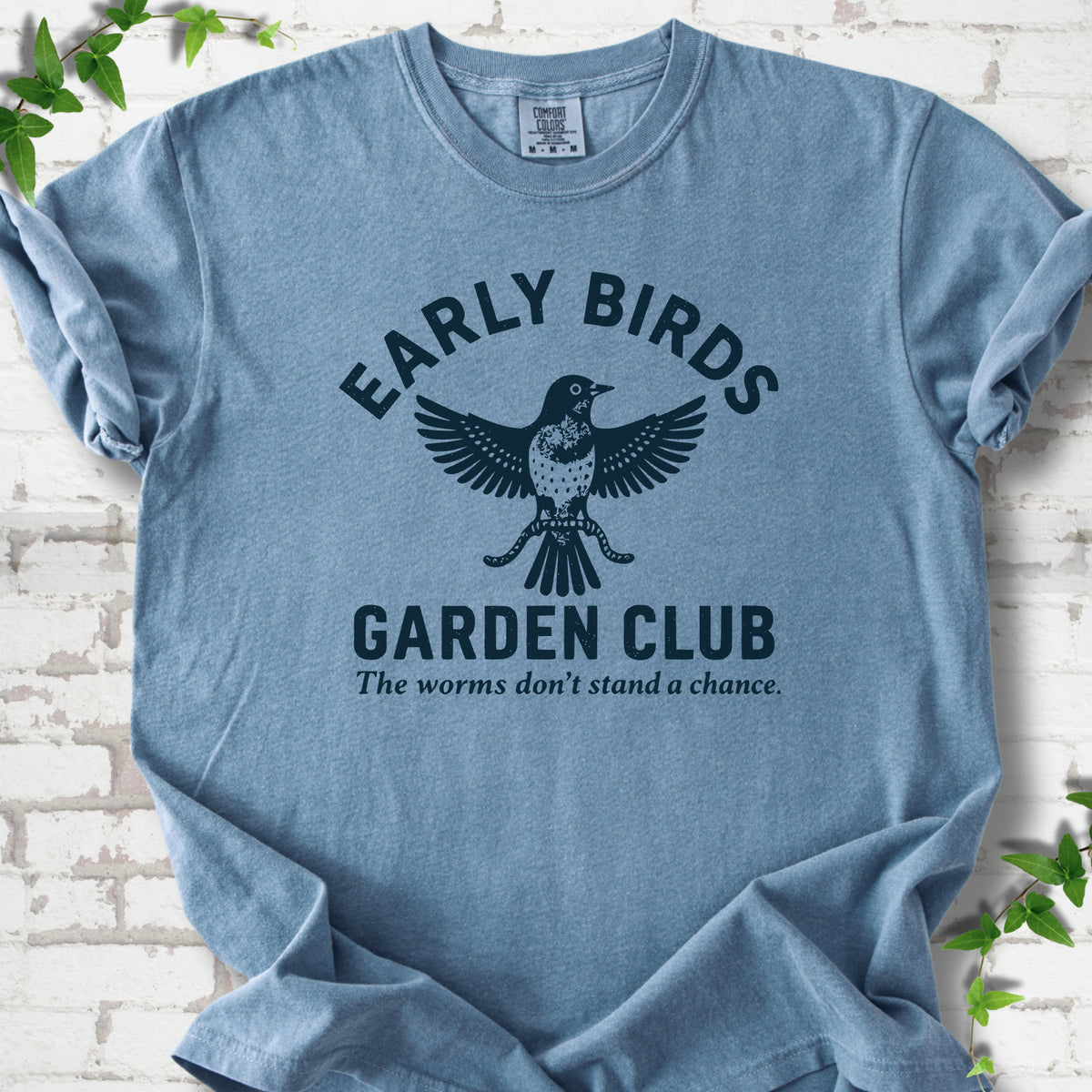 Blue Jean short sleeve T-shirt laid flat on white brick with greenery. Dark navy bird holding worms between "Early Birds" and "Garden Club" chest lettering.