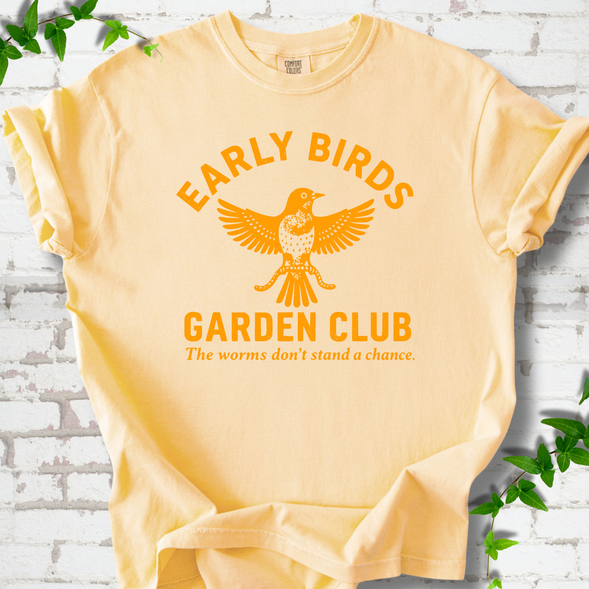 Butter short sleeve T-shirt laid flat on white brick with greenery. Orange bird holding worms between "Early Birds" and "Garden Club" chest lettering.