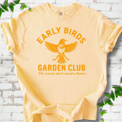 Butter short sleeve T-shirt laid flat on white brick with greenery. Orange bird holding worms between "Early Birds" and "Garden Club" chest lettering.