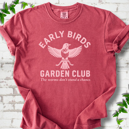 Crimson short sleeve T-shirt laid flat on white brick with greenery. Pale pink bird holding worms between "Early Birds" and "Garden Club" chest lettering.