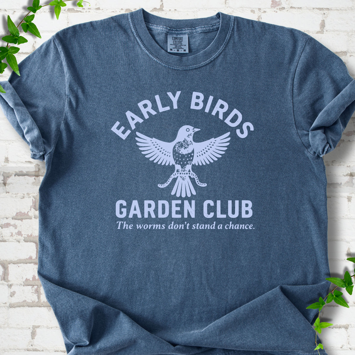 Denim short sleeve T-shirt laid flat on white brick with greenery. Pale blue bird holding worms between "Early Birds" and "Garden Club" chest lettering.