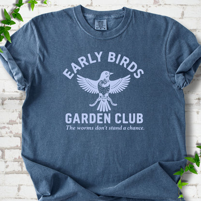 Denim short sleeve T-shirt laid flat on white brick with greenery. Pale blue bird holding worms between "Early Birds" and "Garden Club" chest lettering.