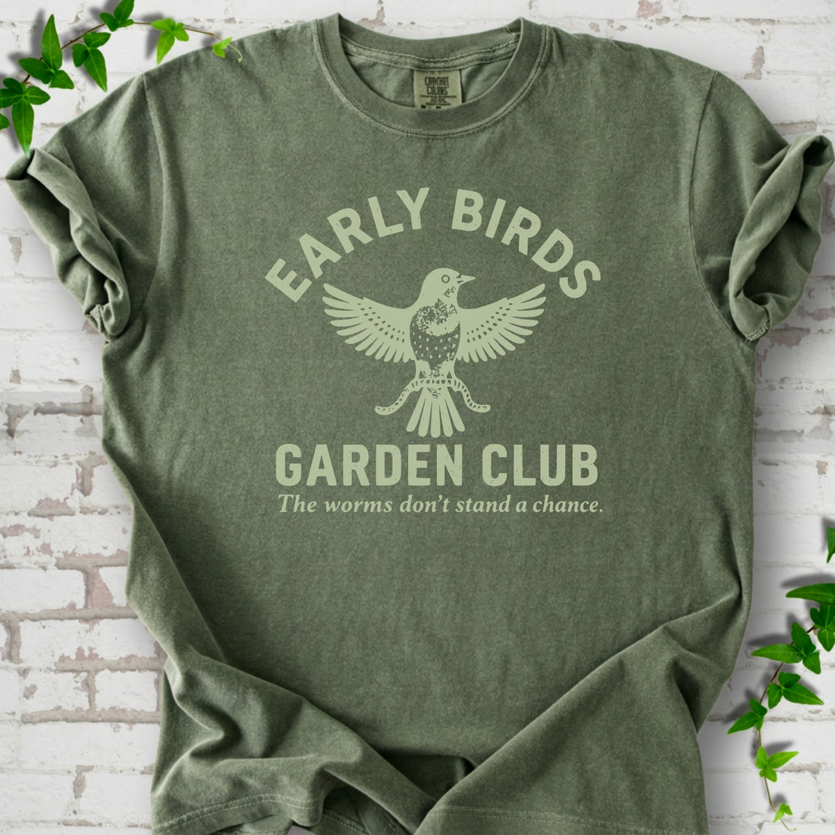 Moss short sleeve T-shirt laid flat on white brick with greenery. Pale green bird holding worms between "Early Birds" and "Garden Club" chest lettering.