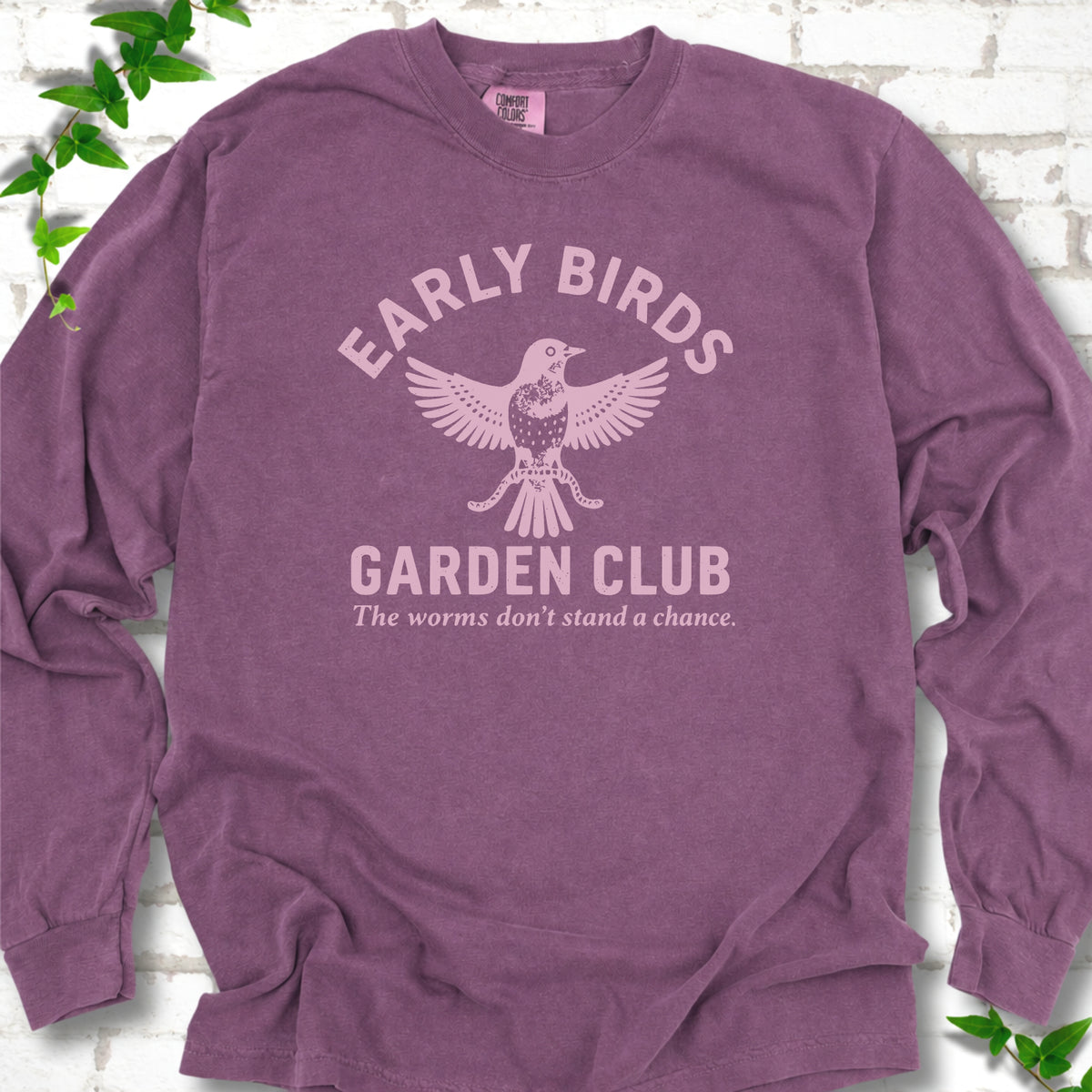 Berry long sleeve T-shirt laid flat on white brick with greenery. Pale pink bird holding worms between "Early Birds" and "Garden Club" chest lettering.