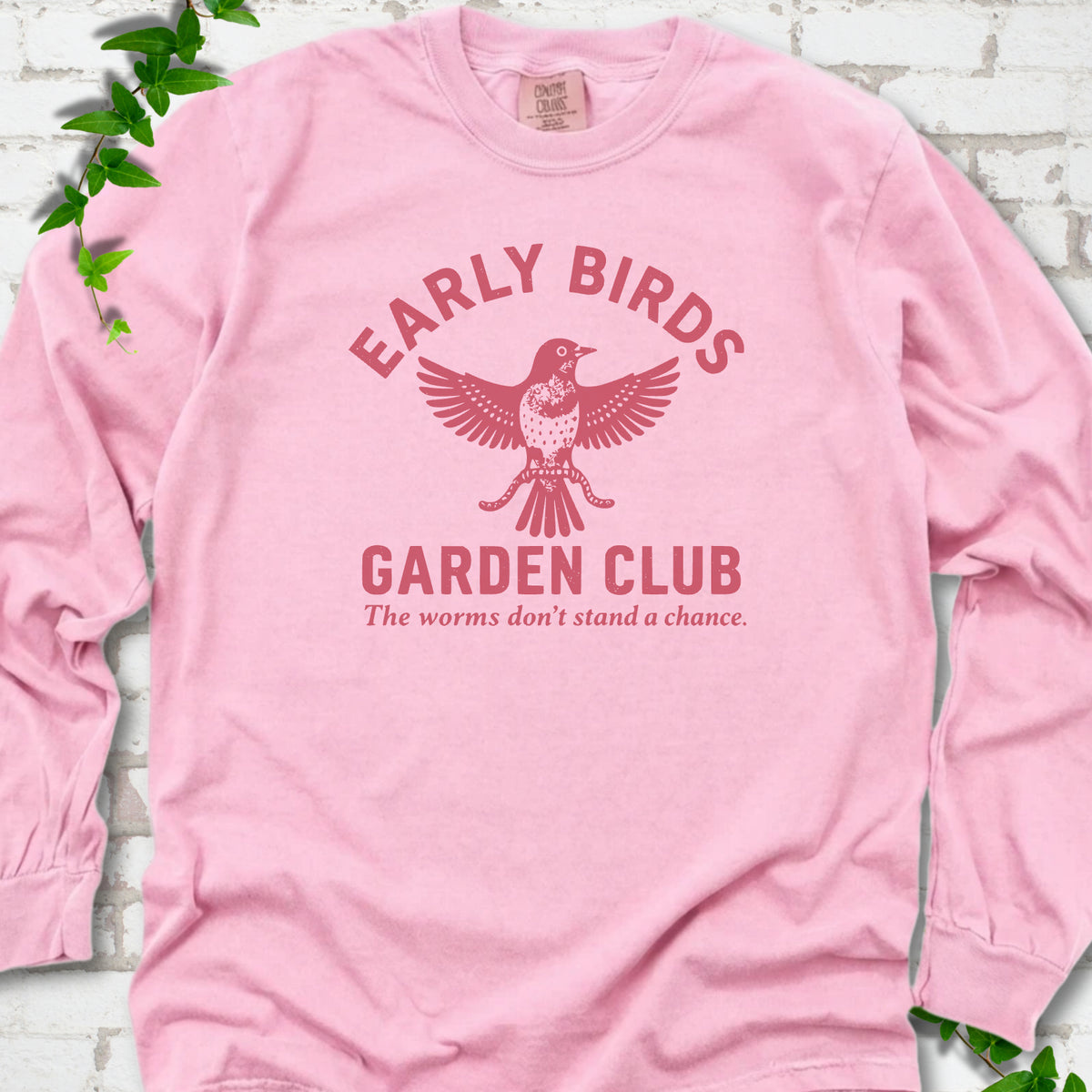 Blossom long sleeve T-shirt laid flat on white brick with greenery. Red bird holding worms between "Early Birds" and "Garden Club" chest lettering.