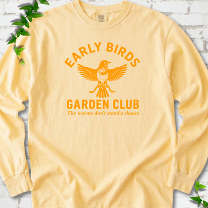 Butter long sleeve T-shirt laid flat on white brick with greenery. Orange bird holding worms between "Early Birds" and "Garden Club" chest lettering.