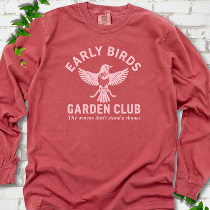 Crimson long sleeve T-shirt laid flat on white brick with greenery. Pale pink bird holding worms between "Early Birds" and "Garden Club" chest lettering.