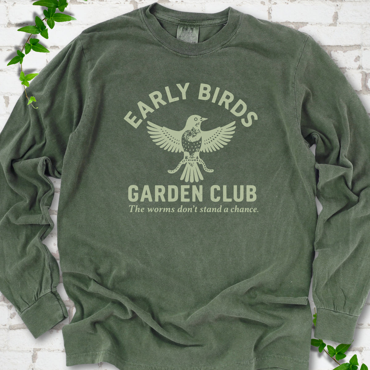 Hemp long sleeve T-shirt laid flat on white brick with greenery. Pale green bird holding worms between "Early Birds" and "Garden Club" chest lettering.