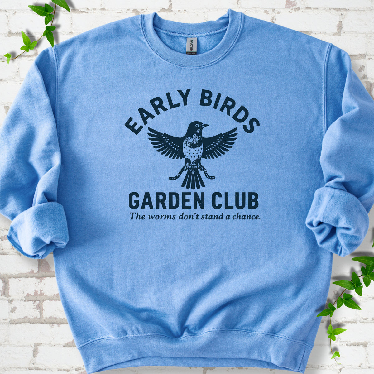 Carolina Blue crewneck sweatshirt laid flat on white brick with greenery. Dark navy bird holding worms between "Early Birds" and "Garden Club" chest lettering.