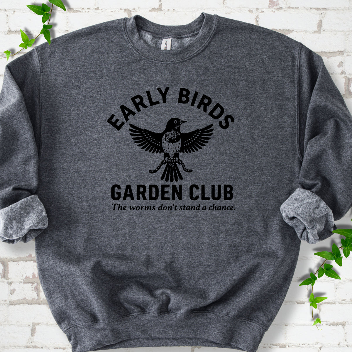Dark Heather crewneck sweatshirt laid flat on white brick with greenery. Black bird holding worms between "Early Birds" and "Garden Club" chest lettering.