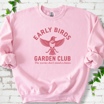 Light Pink crewneck sweatshirt laid flat on white brick with greenery. Red bird holding worms between "Early Birds" and "Garden Club" chest lettering.