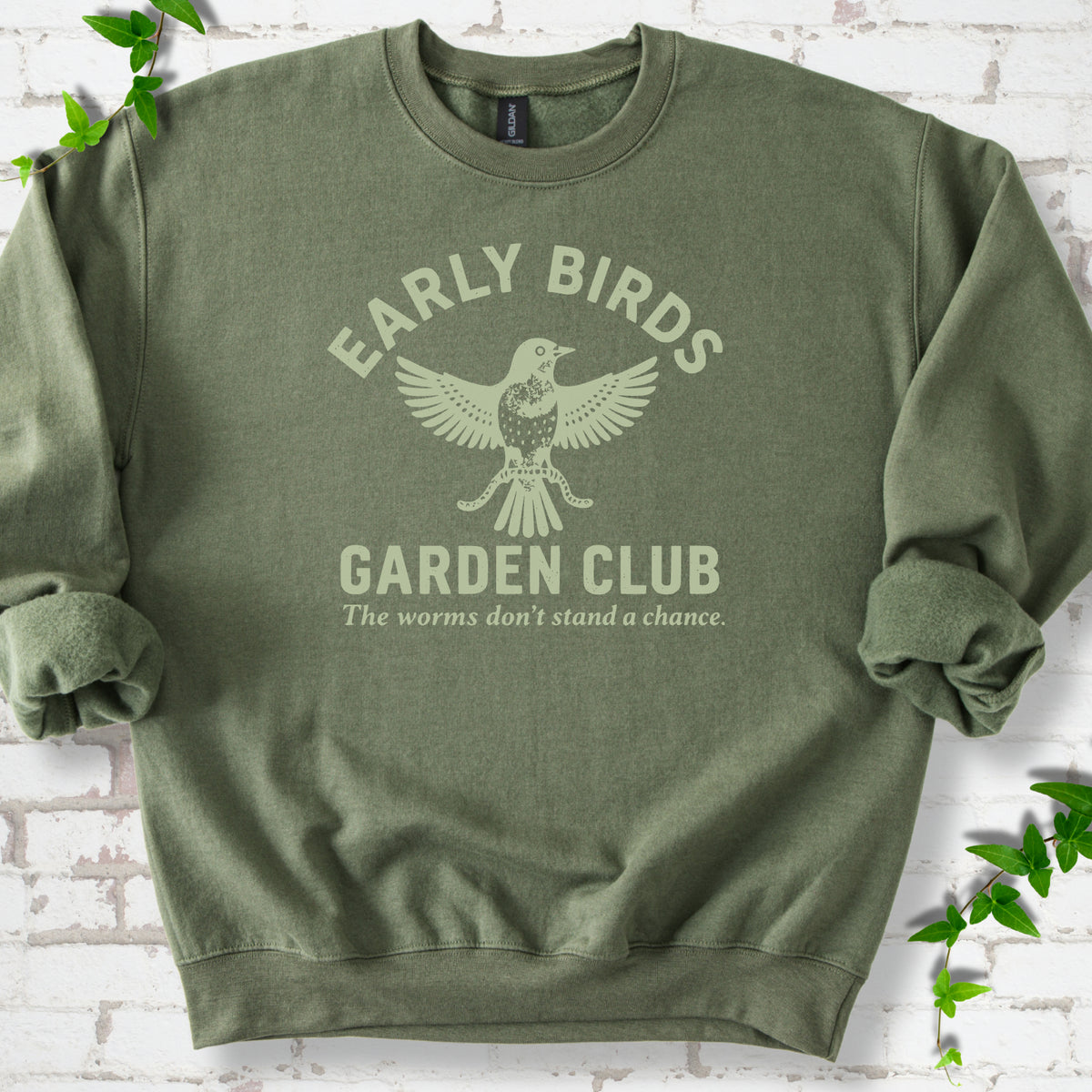 Military Green crewneck sweatshirt laid flat on white brick with greenery. Pale green bird holding worms between "Early Birds" and "Garden Club" chest lettering.
