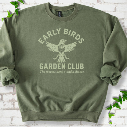 Military Green crewneck sweatshirt laid flat on white brick with greenery. Pale green bird holding worms between "Early Birds" and "Garden Club" chest lettering.